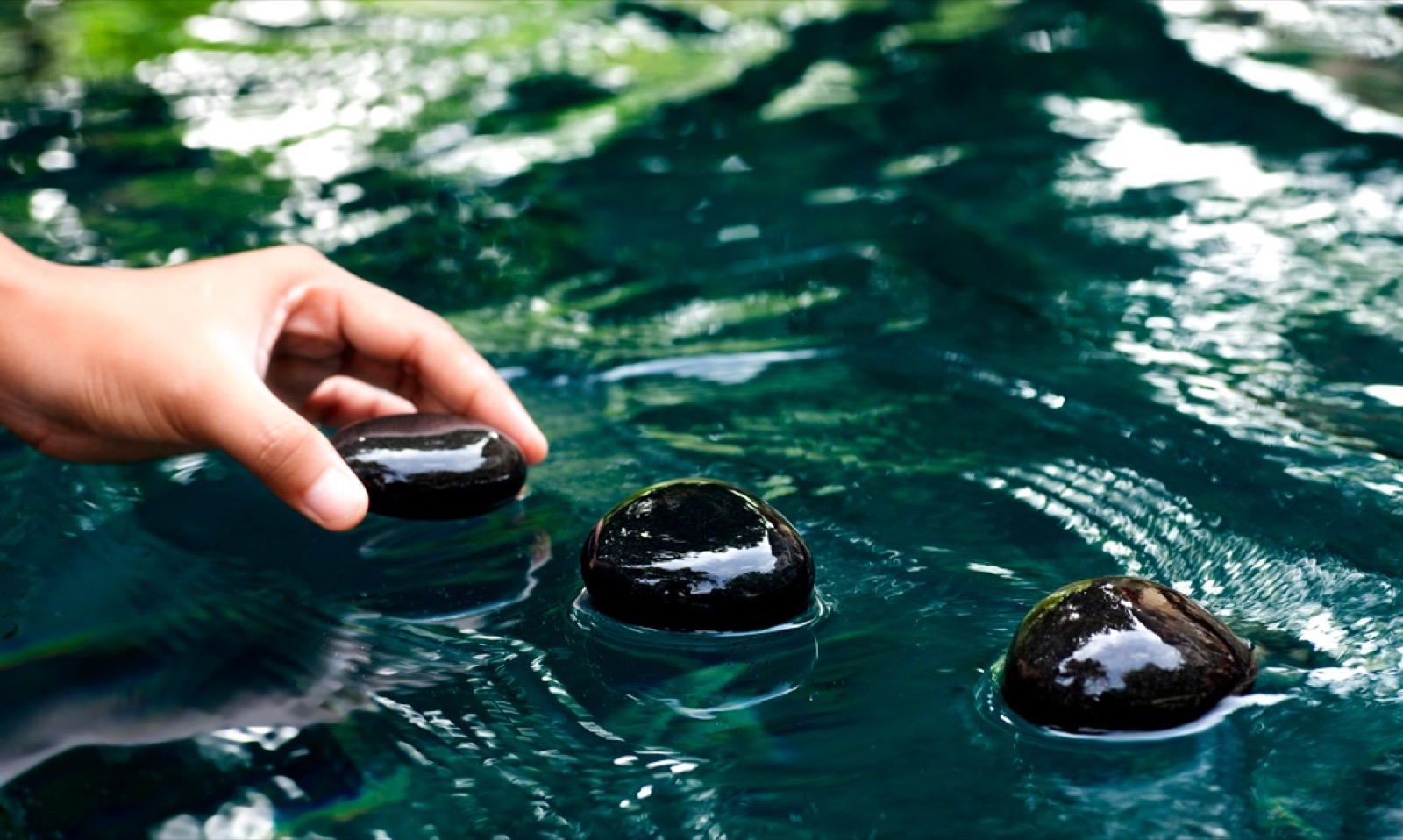 Une main saisit un galet noir poli à la surface d'une eau turquoise, reflets verts