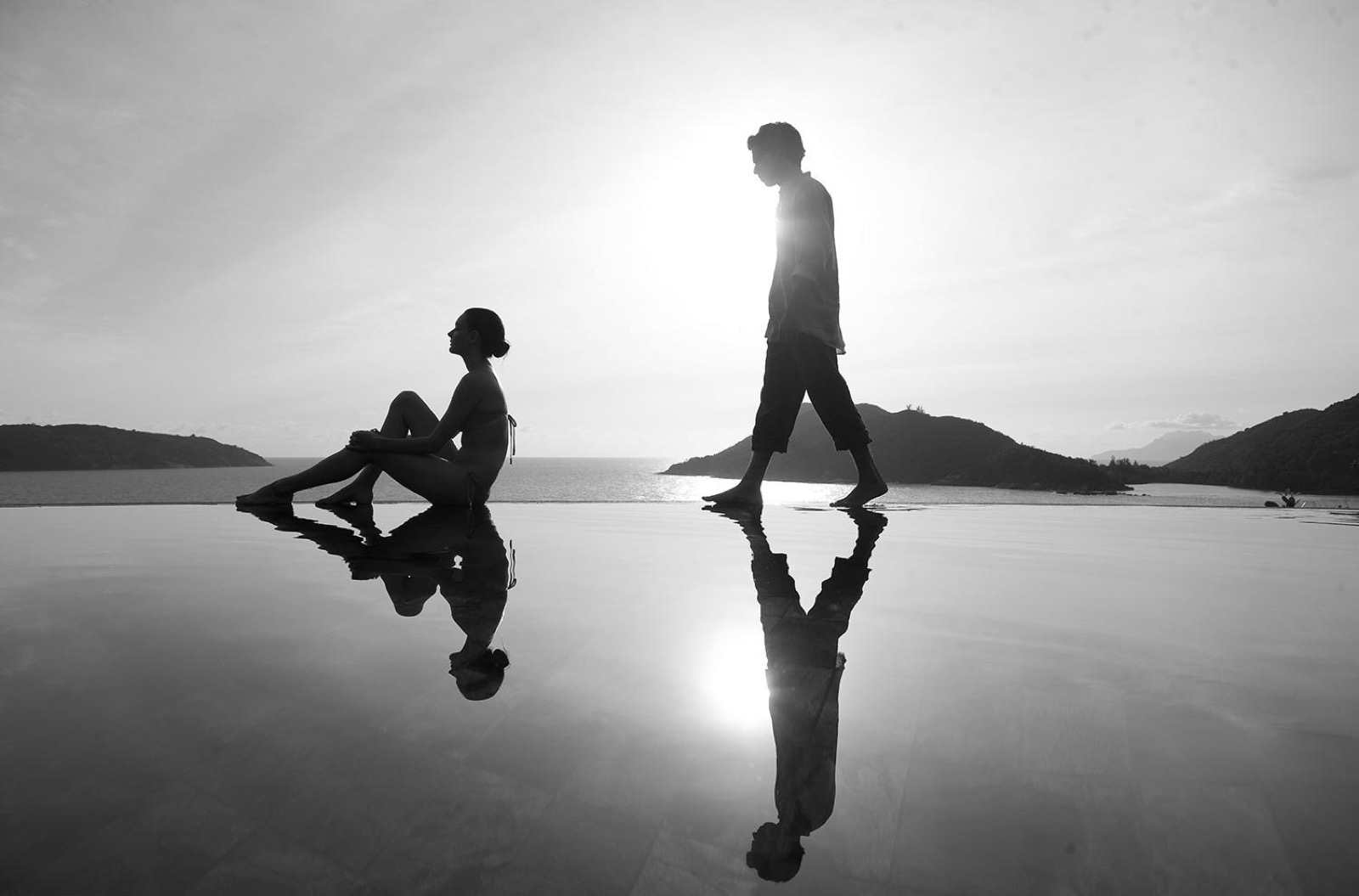 Photo en noir et blanc de deux silhouettes au bord d'une piscine à débordement face à la mer, leurs reflets dessinés dans l'eau immobile