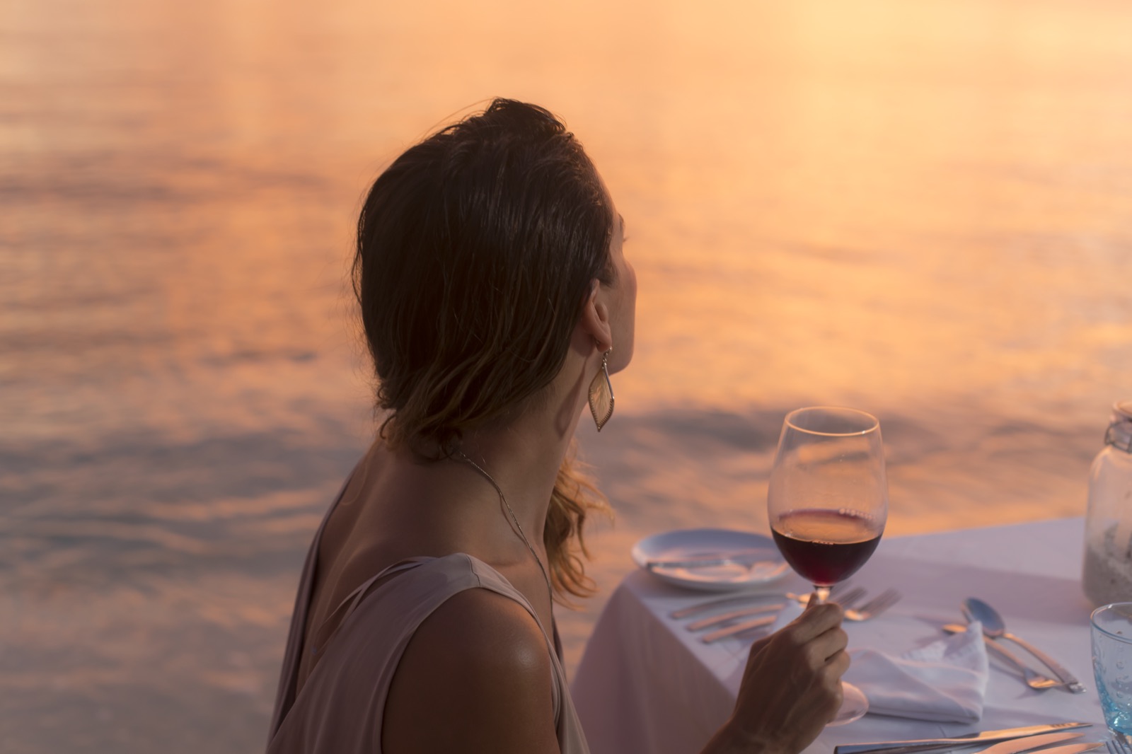 Une femme de dos tient un verre de vin rouge, attablée face à la mer au coucher du soleil, dîner romantique en extérieur