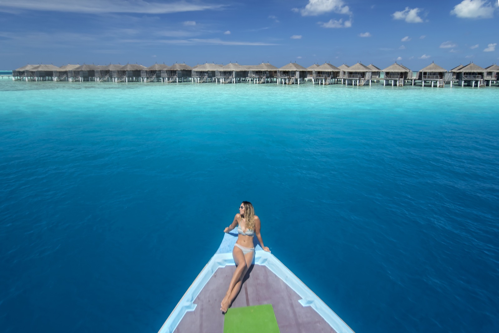 Vue aérienne depuis la proue d'un bateau, une femme allongée face à un alignement de villas sur pilotis au-dessus d'un lagon turquoise