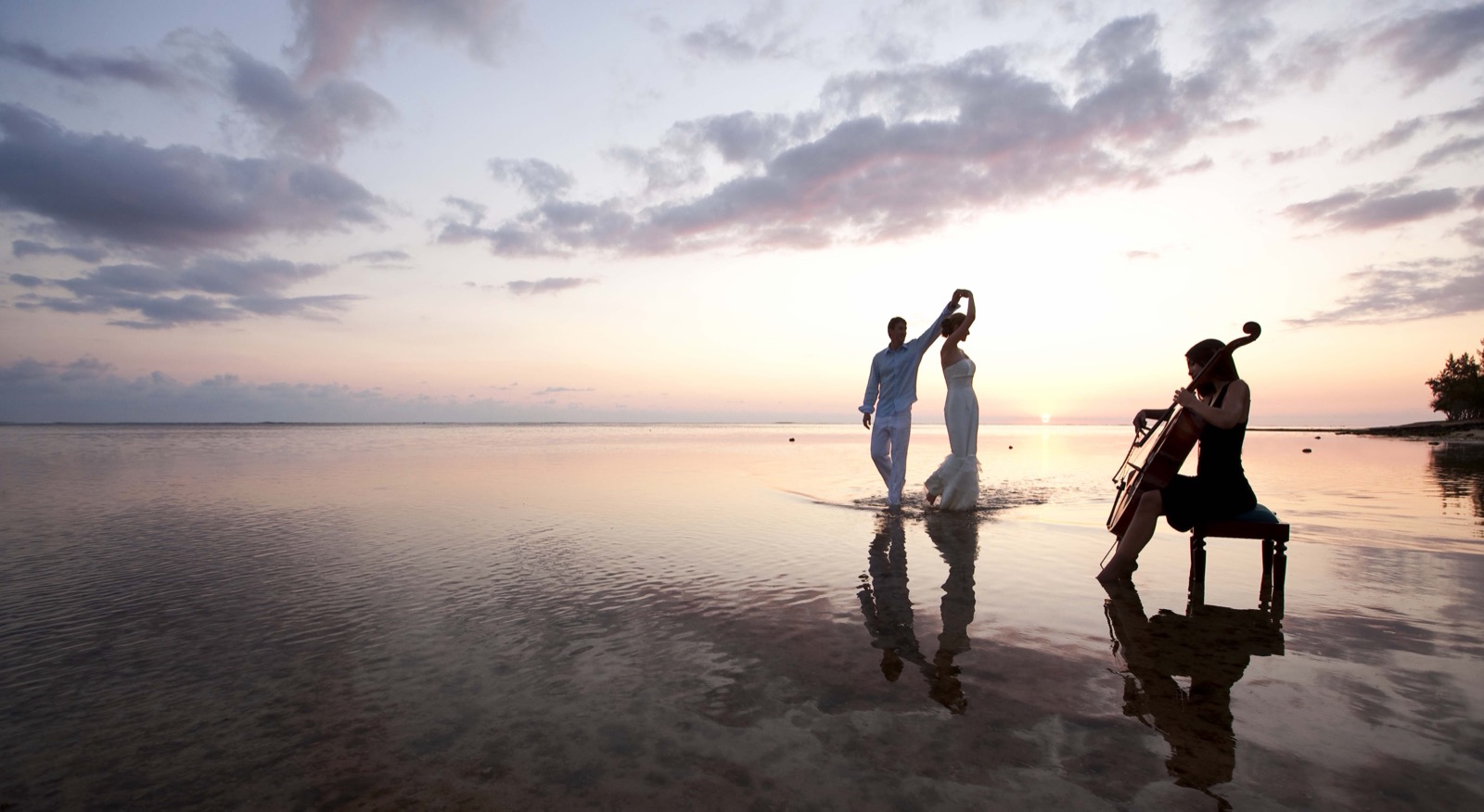 Un couple danse pieds dans l'eau au coucher du soleil tandis qu'une violoncelliste joue pour eux, ciel rose et mauve pastel
