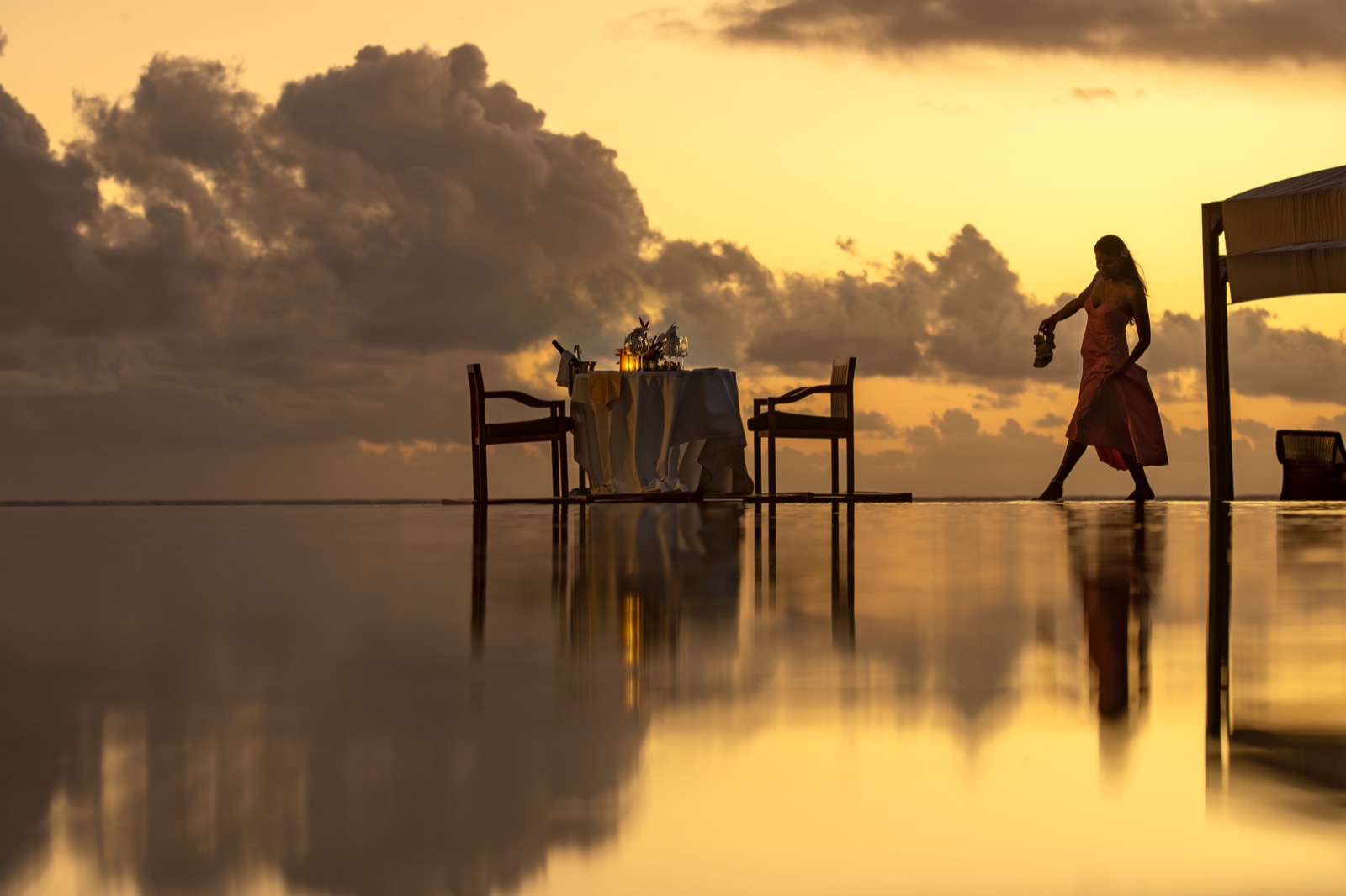 Silhouette d'une femme en robe corail se dirigeant vers une table dressée pour deux sur une surface réfléchissante, lumière dorée de fin de journée