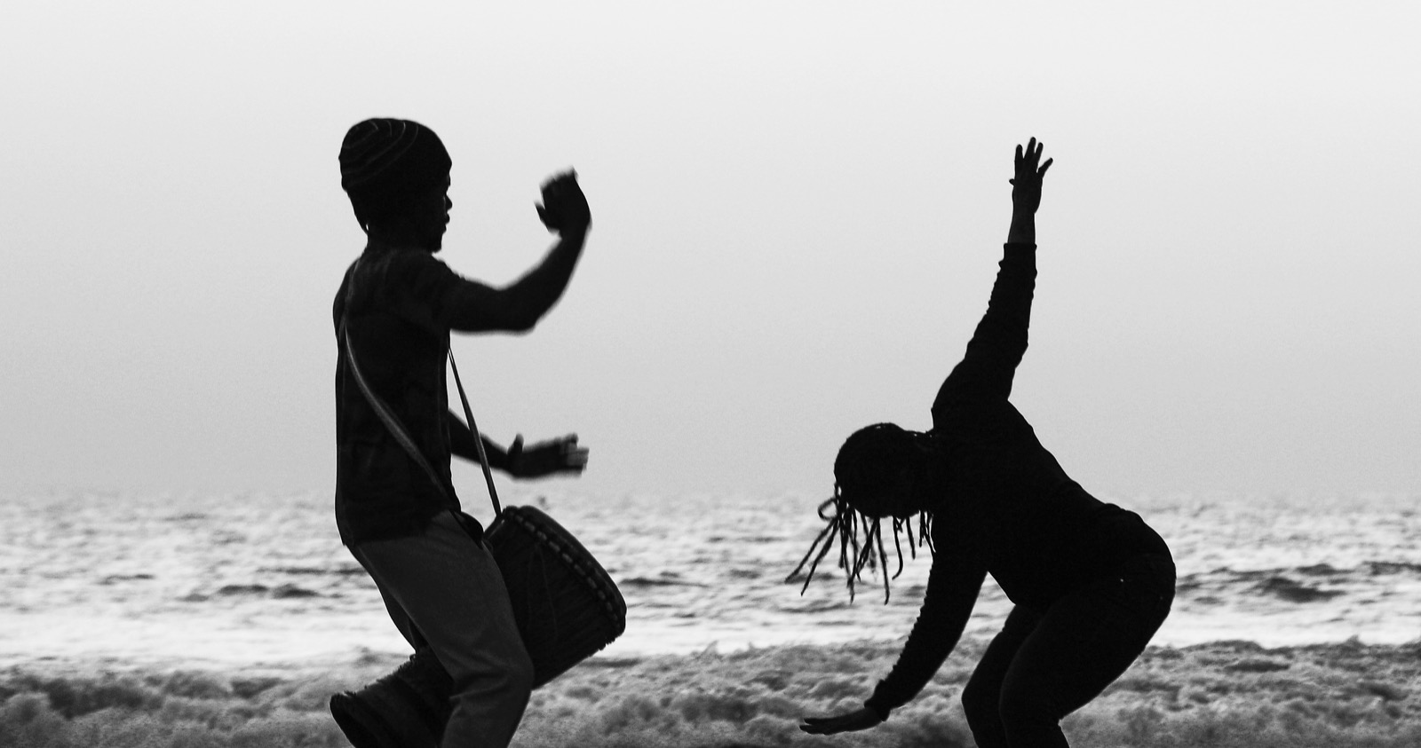 Silhouettes d'un percussionniste et d'une danseuse face à l'océan, photographie en noir et blanc