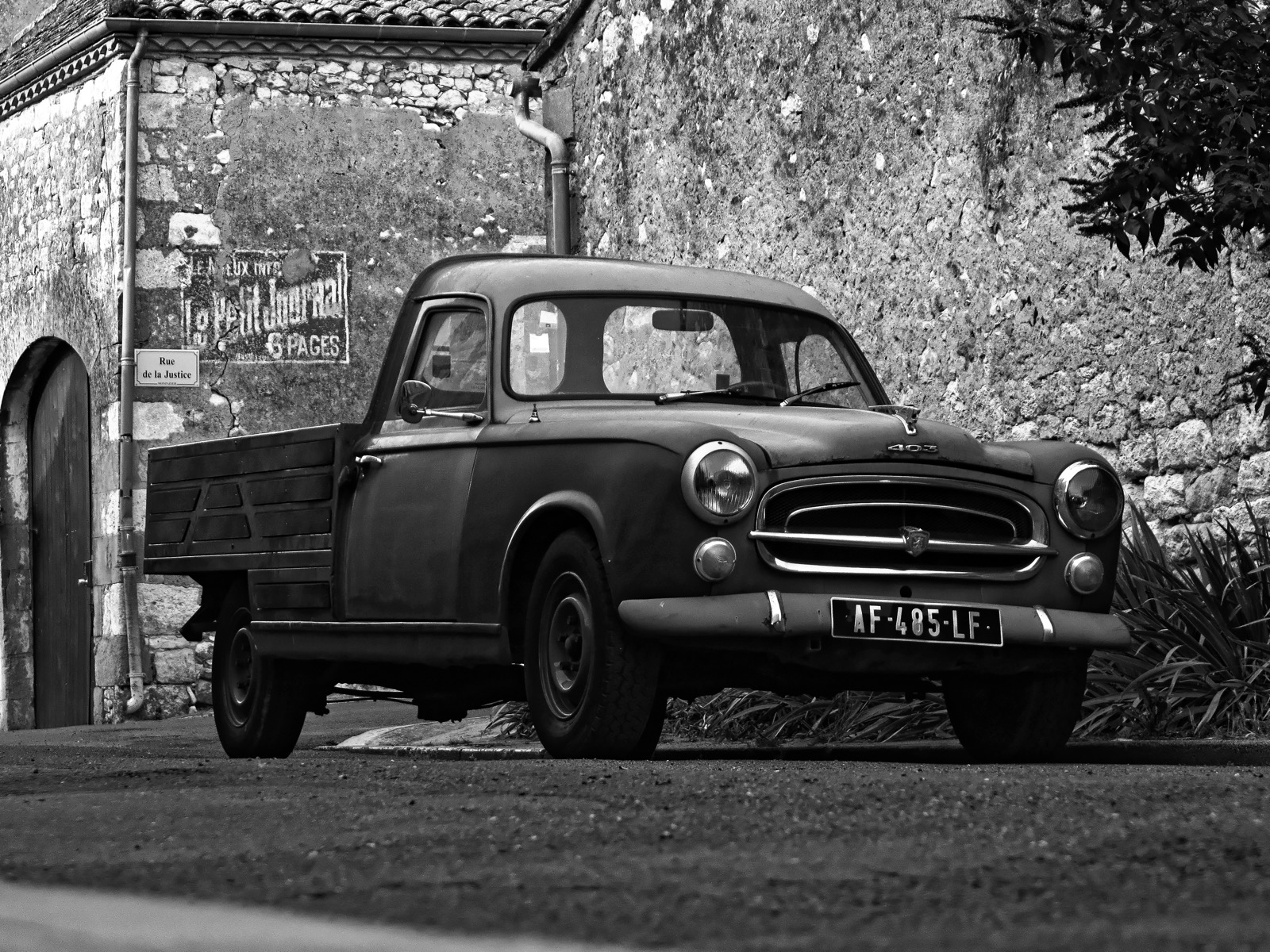 Pickup Peugeot 403 ancien devant un mur de pierre et une réclame peinte, village français, photographie en noir et blanc