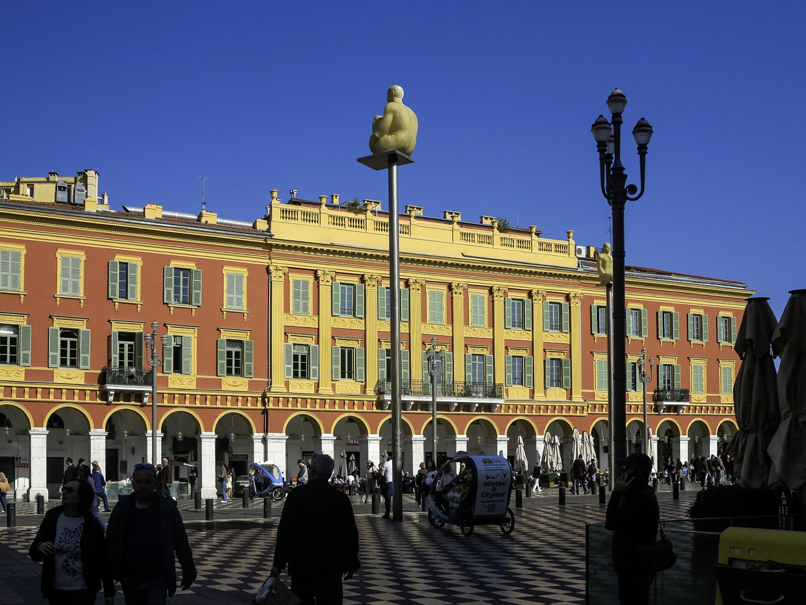 Place Masséna à Nice, façades ocre et rouge sous un ciel bleu, statues de Jaume Plensa sur leurs mâts