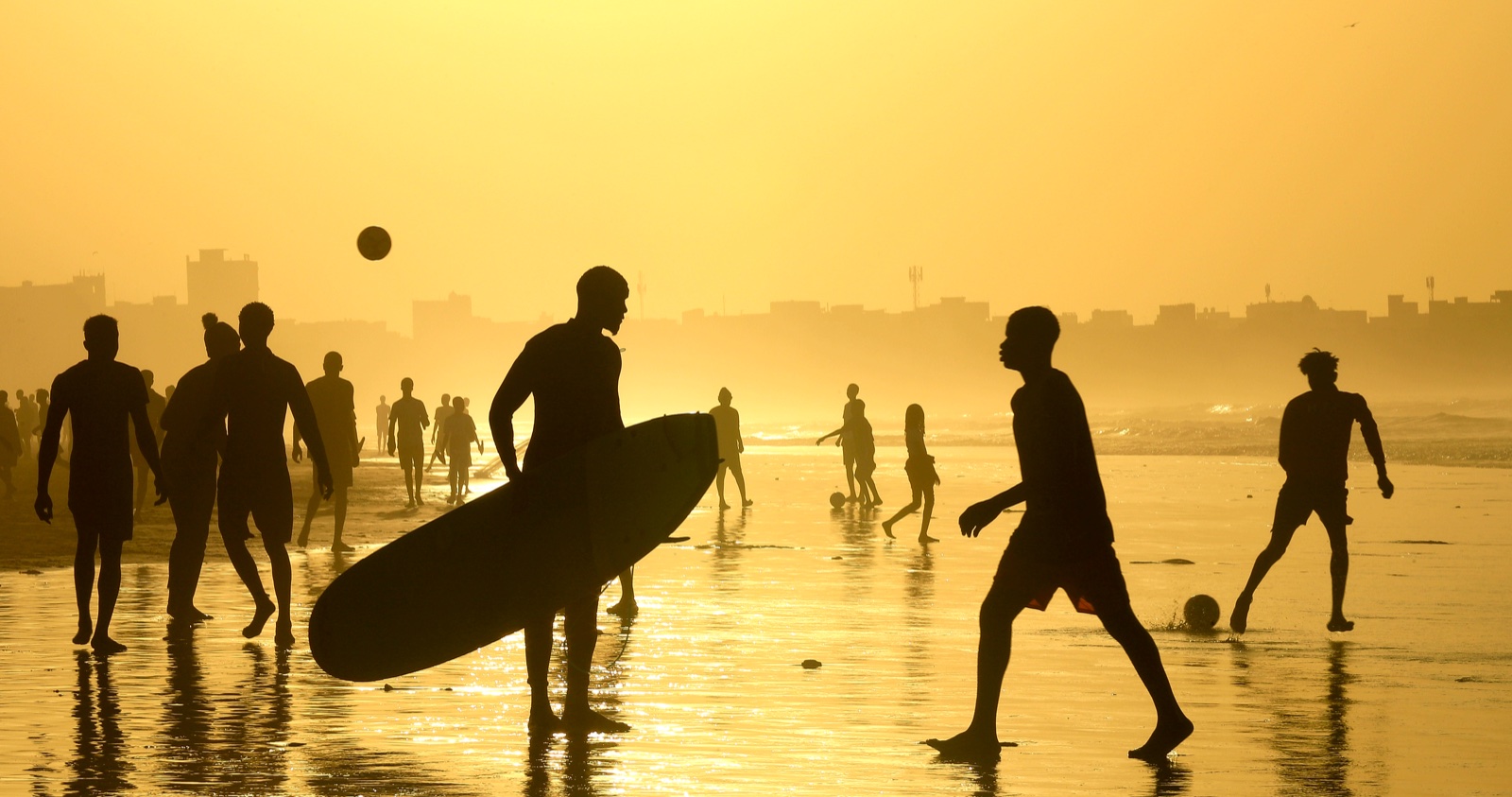 Silhouettes à contre-jour sur une plage dorée au coucher du soleil, un surfeur et des joueurs de football